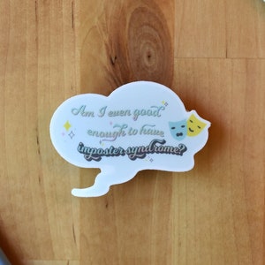May include: White sticker with a speech bubble that says "Am I even good enough to have imposter syndrome?" with two theatrical masks on the right side of the speech bubble. The sticker is on a wooden surface.