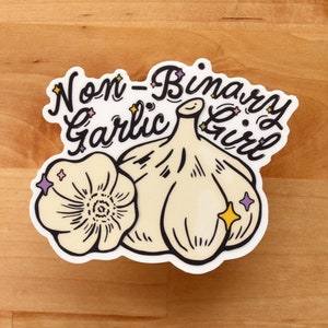 May include: A white sticker with a cartoon illustration of a garlic bulb and a single clove. The text "Non-Binary Garlic Girl" is written in a cursive font above the garlic.