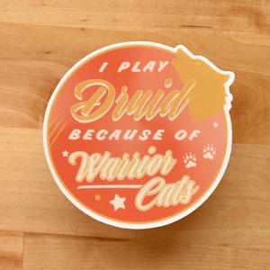 May include: A round sticker with an orange background and white border. The sticker says "I play Druid because of Warrior Cats" with a paw print and a star. The sticker has a silhouette of a cat in the upper right corner.