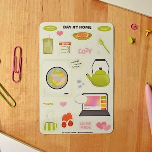 May include: A rectangular sticker sheet titled DAY AT HOME featuring various illustrations including a washing machine, a green kettle, a laptop, a to-do list, a glass of water, a plate of food, slippers, hearts, and the text HOME MADE.