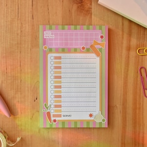 May include: A rectangular notepad with a green and pink striped border, featuring a top grid section labeled PREP AHEAD, a central lined checklist with circular bullet points, and a bottom section labeled SERVE. The design includes small illustrations of a carrot, garlic, and pasta.