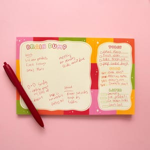 May include: A rectangular paper notepad with a colorful checkered border and a cream-colored center. The notepad features the text BRAIN DUMP, TODAY, SOON, and LATER with handwritten notes. A red ballpoint pen and three paper clips are placed nearby.