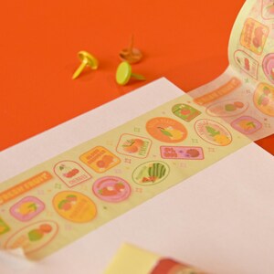 A roll of washi tape is rolled onto a white paper pad on top of a red background. The washi tape is a wide 30mm width featuring artwork of fruit stickers. The tape's label reads "Fruit Stickers Wide Washi Tape By Franki Hanke 30mm by 10M."