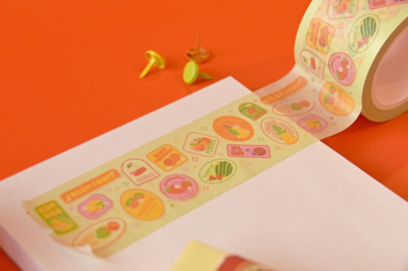 A roll of washi tape is rolled onto a white paper pad on top of a red background. The washi tape is a wide 30mm width featuring artwork of fruit stickers. The tape's label reads "Fruit Stickers Wide Washi Tape By Franki Hanke 30mm by 10M."