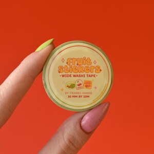 A woman's hand holds a roll of washi tape in front of a red background. The washi tape is a wide 30mm width featuring artwork of fruit stickers. The tape's label reads "Fruit Stickers Wide Washi Tape By Franki Hanke 30mm by 10M."