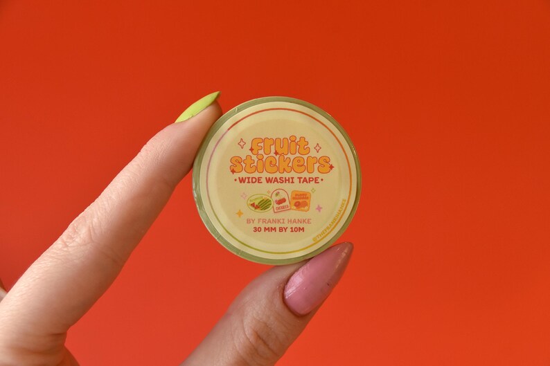 A woman's hand holds a roll of washi tape in front of a red background. The washi tape is a wide 30mm width featuring artwork of fruit stickers. The tape's label reads "Fruit Stickers Wide Washi Tape By Franki Hanke 30mm by 10M."
