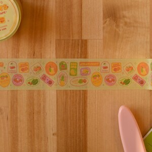 A roll of washi tape is rolled out over a wooden desktop. The washi tape is a wide 30mm width featuring artwork of fruit stickers. The tape's label reads "Fruit Stickers Wide Washi Tape By Franki Hanke 30mm by 10M."