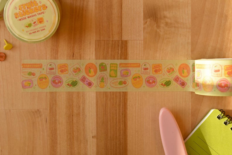 A roll of washi tape is rolled out over a wooden desktop. The washi tape is a wide 30mm width featuring artwork of fruit stickers. The tape's label reads "Fruit Stickers Wide Washi Tape By Franki Hanke 30mm by 10M."