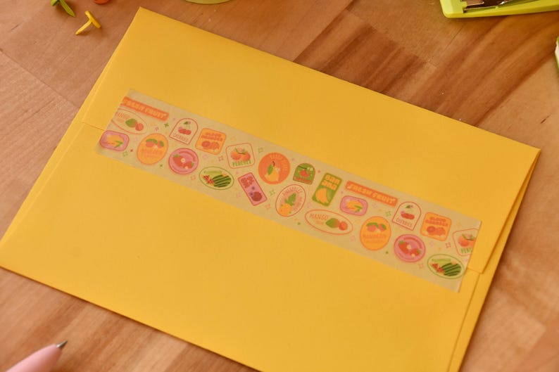 A yellow envelope is sealed with a piece of washi tape. The washi tape is a wide 30mm width featuring artwork of fruit stickers. The tape's label reads "Fruit Stickers Wide Washi Tape By Franki Hanke 30mm by 10M."