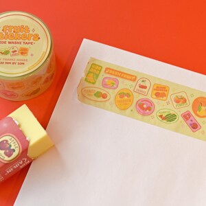A roll of washi tape is rolled onto a white piece of paper. The washi tape is a wide 30mm width featuring artwork of fruit stickers. The tape's label reads "Fruit Stickers Wide Washi Tape By Franki Hanke 30mm by 10M."