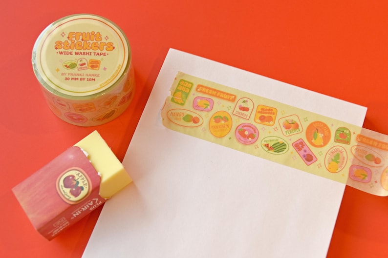 A roll of washi tape is rolled onto a white piece of paper. The washi tape is a wide 30mm width featuring artwork of fruit stickers. The tape's label reads "Fruit Stickers Wide Washi Tape By Franki Hanke 30mm by 10M."