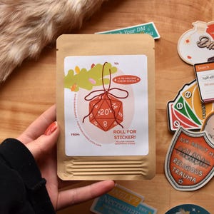 May include: A brown paper package with a white label featuring a red D20 die graphic and the text "Roll for Sticker!". Various stickers with text and graphics are scattered around the package.