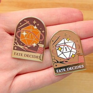May include: Two gold-toned enamel pins with a red and orange thread design. Each pin features a die with a single dot and the text "Fate Decides".