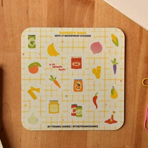 May include: A square sticker sheet with a yellow grid pattern featuring colorful illustrations of fruits, vegetables, and jars. Text at the top reads "Grocery Haul with 17 Waterproof Stickers." Includes a banana, onion, carrot, and more.