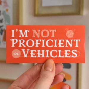 May include: A red sticker with white text that reads "I'm not proficient in vehicles". A white circle with the word "in" is in the upper left corner of the sticker. A white dice with a black outline is in the upper right corner of the sticker.