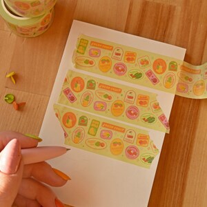 Washi tape has been repeatedly swatched on a white paper pad. A hand hold a pink pen poised to write. The washi tape is a wide 30mm width featuring artwork of fruit stickers. The tape's label reads "Fruit Stickers Wide Washi Tape By Franki Hanke 30mm by 10M."