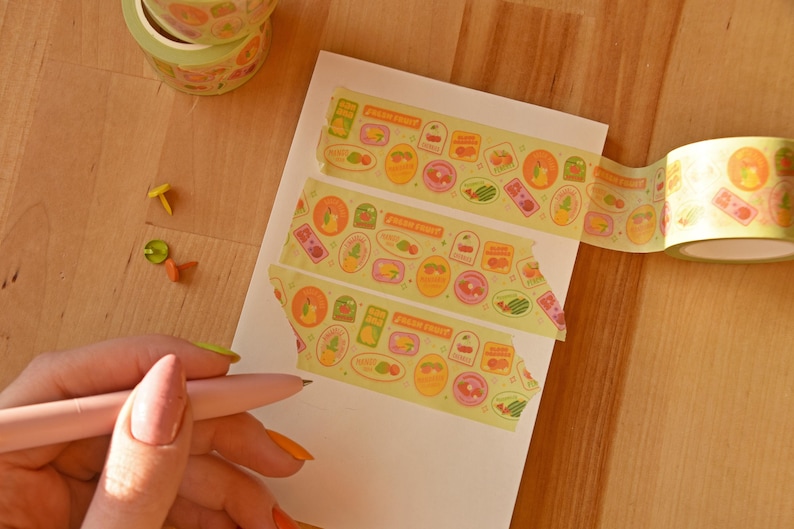 Washi tape has been repeatedly swatched on a white paper pad. A hand hold a pink pen poised to write. The washi tape is a wide 30mm width featuring artwork of fruit stickers. The tape's label reads "Fruit Stickers Wide Washi Tape By Franki Hanke 30mm by 10M."