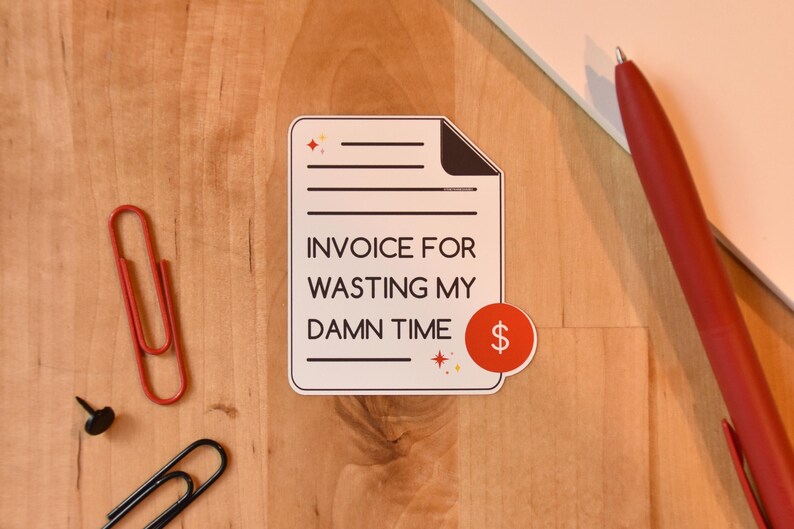 Op de afbeelding: Een sticker met de tekst "INVOICE FOR WASTING MY DAMN TIME" en een dollarteken. Een rode pen, een rode paperclip, een zwarte paperclip en een zwarte punaise zijn ook zichtbaar op een houten oppervlak.
