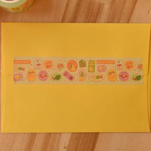 A yellow envelope is sealed with a piece of washi tape. The washi tape is a wide 30mm width featuring artwork of fruit stickers. The tape's label reads "Fruit Stickers Wide Washi Tape By Franki Hanke 30mm by 10M."