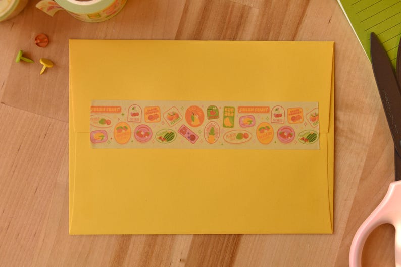 A yellow envelope is sealed with a piece of washi tape. The washi tape is a wide 30mm width featuring artwork of fruit stickers. The tape's label reads "Fruit Stickers Wide Washi Tape By Franki Hanke 30mm by 10M."