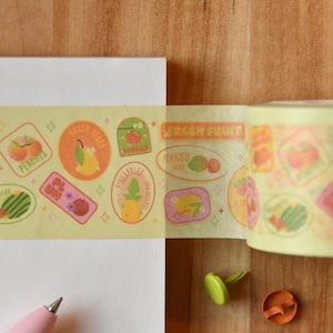 A roll of washi tape is rolled onto a white piece of paper alongside a pink pen. The washi tape is a wide 30mm width featuring artwork of fruit stickers. The tape's label reads "Fruit Stickers Wide Washi Tape By Franki Hanke 30mm by 10M."