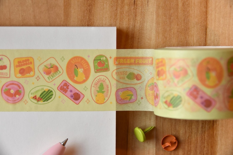 A roll of washi tape is rolled onto a white piece of paper alongside a pink pen. The washi tape is a wide 30mm width featuring artwork of fruit stickers. The tape's label reads "Fruit Stickers Wide Washi Tape By Franki Hanke 30mm by 10M."