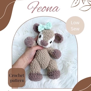 Pode incluir: Um peluche de veado de crochê chamado Feona, em tons de castanho, branco e um laço verde menta. A imagem inclui o texto "Low Sew" e "Crochet pattern". O peluche é segurado por uma mão.