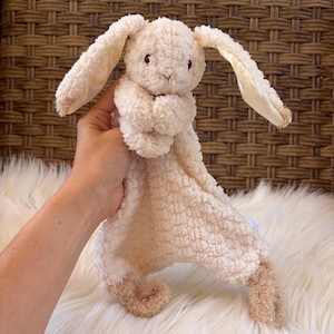 May include: A cream-colored plush security blanket featuring a rabbit head with long, floppy ears. The rabbit has embroidered facial features and a soft, textured body that transitions into a flat, square-shaped blanket with tan-colored feet.