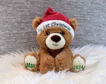 May include: A brown teddy bear wearing a red and white Santa hat with the text "My 1st Christmas". The bear has the name "Max" and the year "2024" embroidered on its paws. The bear is sitting on a white, fluffy surface.
