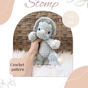 May include: A light blue crochet dinosaur toy with white detailing. The word "Stomp" is written above the dinosaur. The words "No Sew" and "Crochet pattern" are also visible. The toy is displayed on a white, fluffy surface.