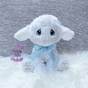 May include: A white plush lamb with blue eyes and a blue bow. The lamb is sitting on a white fluffy surface. The lamb has embroidered text on its paws that says "God Bless Mateo" and "Baptized 6.27.21".