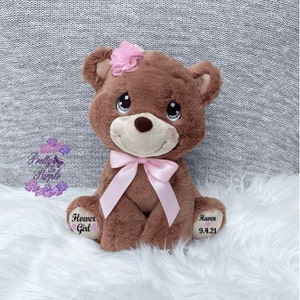 May include: A brown plush teddy bear with a pink bow and a pink flower on its head. The bear has embroidered text on its paws that reads "Haven 9.4.21" and "Flower Girl".