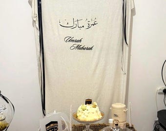 May include: A beige fabric backdrop featuring the text Umrah Mubarak in black calligraphy and English script, hung with black ribbons. Below, a table displays a white frosted cake, a small Kaaba-themed sign, and a beverage dispenser.