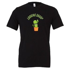 May include: Black t-shirt featuring a cartoon cactus wearing sunglasses in an orange pot. The text "LOOKING SHARP" arches above the cactus in green. The shirt is a classic crew neck style.