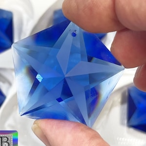 Può includere: Un ciondolo a forma di stella in cristallo blu con un foro nella parte superiore per appenderlo. Il ciondolo è sfaccettato e ha una finitura trasparente e iridescente.