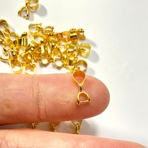 Może przedstawiać: Stosik złotych metalowych zapięć do biżuterii, niektóre leżą na dłoni, inne są rozrzucone na białym tle.