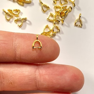 Puede incluir: Primer plano de una mano sosteniendo una anilla de colgante de metal dorado. La anilla tiene forma de corazón con un pequeño bucle en la parte superior. Hay muchas otras anillas similares esparcidas sobre una superficie blanca.