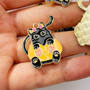 May include: A set of five gold-tone metal charms featuring black cats in various poses. The charms are decorated with orange and pink enamel and have a textured design. The charms are perfect for adding a touch of whimsy to jewelry, crafts, and other projects.