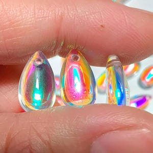以下が含まれることがあります： 透明で虹色のしずく型のビーズで、上部に穴があります。ビーズはピンク、黄色、青など、虹色の色を反射します。白い表面にいくつかのビーズが散らばっており、3つが指の間に挟まれています。