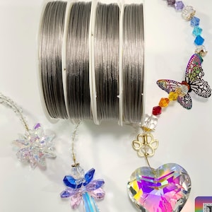 Może przedstawiać: Trzy szpulki srebrnego drutu jubilerskiego, każda o innej grubości. Drut jest używany do tworzenia biżuterii, łapaczy snów i innych rękodzieł. Obraz przedstawia również różne kryształowe zawieszki, w tym serce, anioła i motyla.