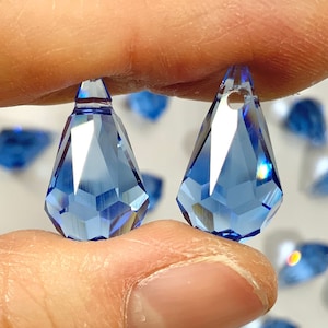 Może przedstawiać: Zbliżenie na dłoń trzymającą dwa jasnoniebieskie kryształowe wisiorek w kształcie łzy. Wisiorek ma fasetowany wzór i mały otwór u góry do przewleczenia sznurka. Na powierzchni poniżej rozrzuconych jest wiele innych wisiorów.