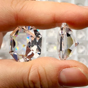May include: Two clear octagon-shaped crystal rhinestones are held in a hand. The rhinestones are faceted and have a rainbow effect. The rhinestones are on a background of many other rhinestones in a grid.