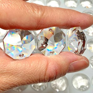 May include: Three clear, faceted crystal pendants held between fingers. The pendants are octagonal with multiple facets, reflecting light. The background features many similar crystal pieces.