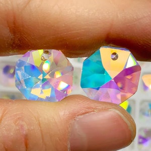 May include: Two iridescent clear glass octagon shaped pendants with a hole at the top for stringing. The pendants are held in a hand against a background of many similar pendants.