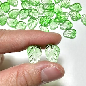 May include: A collection of small, translucent light green glass beads shaped like leaves. Each bead features a detailed vein pattern and a single hole at the top for stringing. The beads are scattered across a plain surface with two held up.