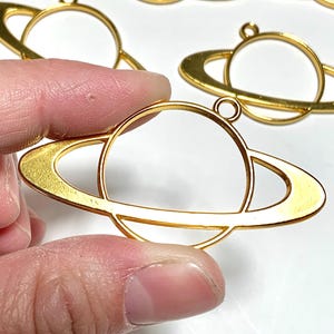 Peut inclure: Charm en métal doré en forme de planète. Les breloques présentent un centre circulaire avec un anneau autour, ressemblant à la planète Saturne. Chaque breloque a une petite boucle en haut pour l'attacher à des bijoux ou à d'autres créations. Plusieurs breloques sont visibles.