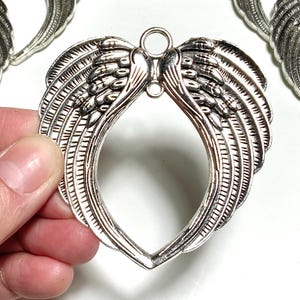Op de afbeelding: Zilverkleurige metalen engelenvleugelbedels. De bedels zijn gedetailleerd met veertjes en hebben een lusje om op te hangen. Er zijn meerdere bedels zichtbaar, waarvan er één in een hand wordt gehouden. Ideaal voor het maken van sieraden of knutselprojecten.