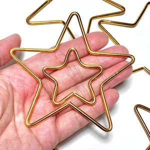 Puede incluir: Formas de estrella de alambre dorado, algunas anidadas, sobre un fondo blanco. Las estrellas varían en tamaño, algunas se sostienen en una mano. El alambre es delgado y parece ser de metal. Las estrellas son de color dorado brillante.