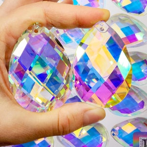 May include: Two iridescent crystal teardrop pendants with a faceted design. The pendants have a hole at the top for hanging. The pendants are held in a hand against a background of many other similar pendants.