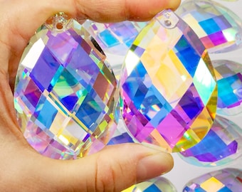 Op de afbeelding: Twee regenboogkleurige kristallen druppelhangers met een facetgeslepen ontwerp. De hangers hebben een gat aan de bovenkant om op te hangen. De hangers worden in een hand gehouden tegen een achtergrond van veel andere vergelijkbare hangers.